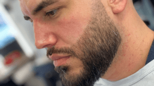 Close-up of a mans face in profile, showcasing a well-groomed beard and focused expression, as if he just stepped out of the finest barbershop in Bryan, TX. Background is artfully blurred.