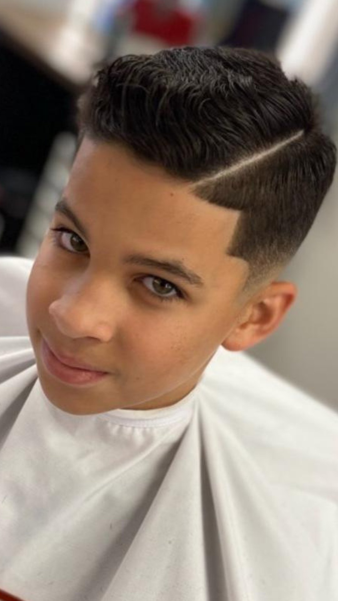 A boy with a short, styled haircut and a side part, draped in a white barber cape, looks towards the camera at Brayn Barber Shop.