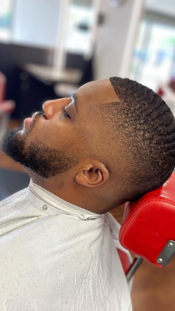 A person with a neatly trimmed beard and short, well-groomed hair sits in a red barbershop chair at a barber shop in Bryan, Texas. Theyre wearing a white cape, leaned back, showcasing their stylish haircut and relaxed posture.