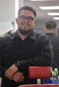 A person with glasses and a beard stands smiling, leaning on a red chair in a room with bright lighting.